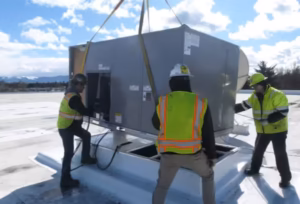 Crane lifting a rooftop HVAC unit while installers in safety vests guide it
