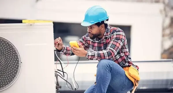 Technician in hard hat testing an outdoor AC unit with a multimeter
